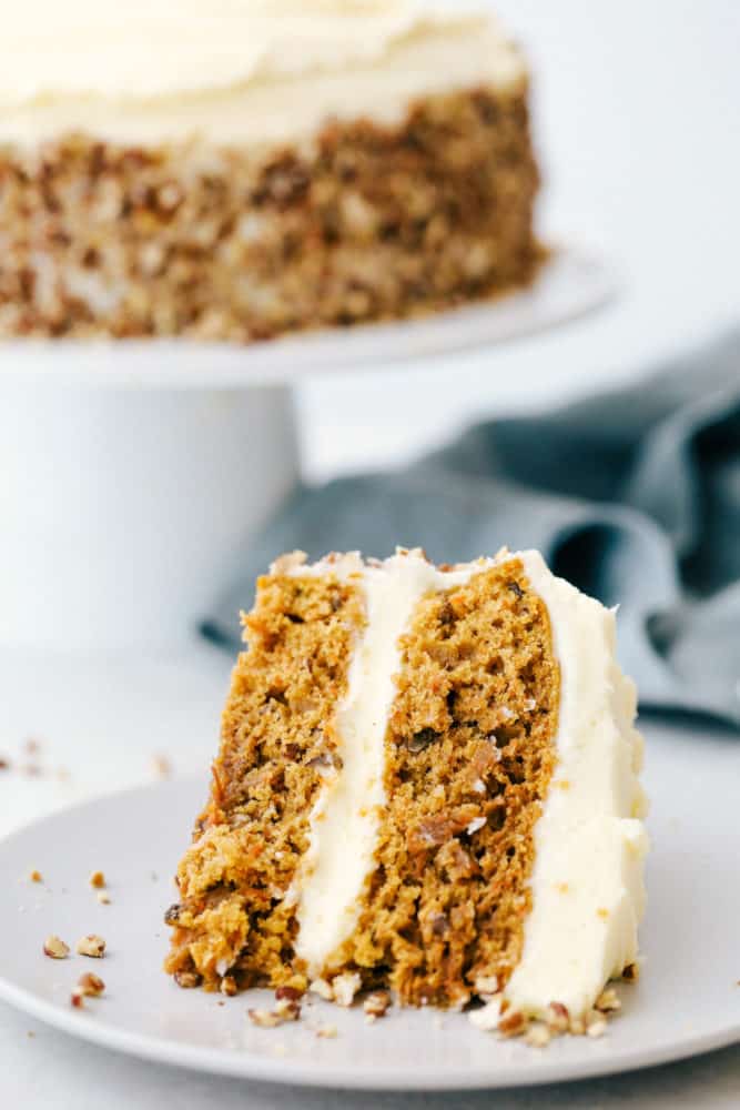 Une tranche de gâteau aux carottes sur une assiette avec le gâteau aux carottes en arrière-plan sur un plateau à gâteaux. 