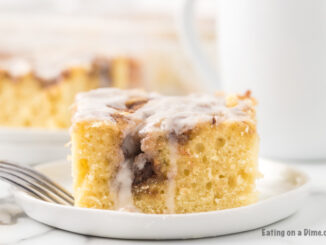 A piece of the cinnamon roll cake on a white plate with a fork with the rest of the cake in the back ground.