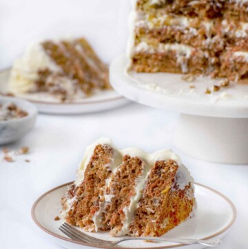 tranche de gâteau aux carottes sur une assiette avec une fourchette avec une autre tranche et le reste du gâteau entier sur un piédestal en arrière-plan