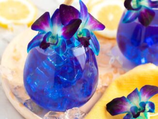2 blue drinks in round glasses sitting on a light wooden board with a yellow cloth, garnished with purple and blue orchids